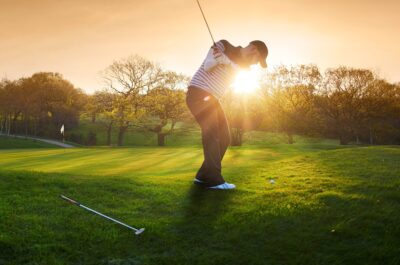 a lone golfer chips onto the green , the low sun is coming from behind him and flaring to camera.